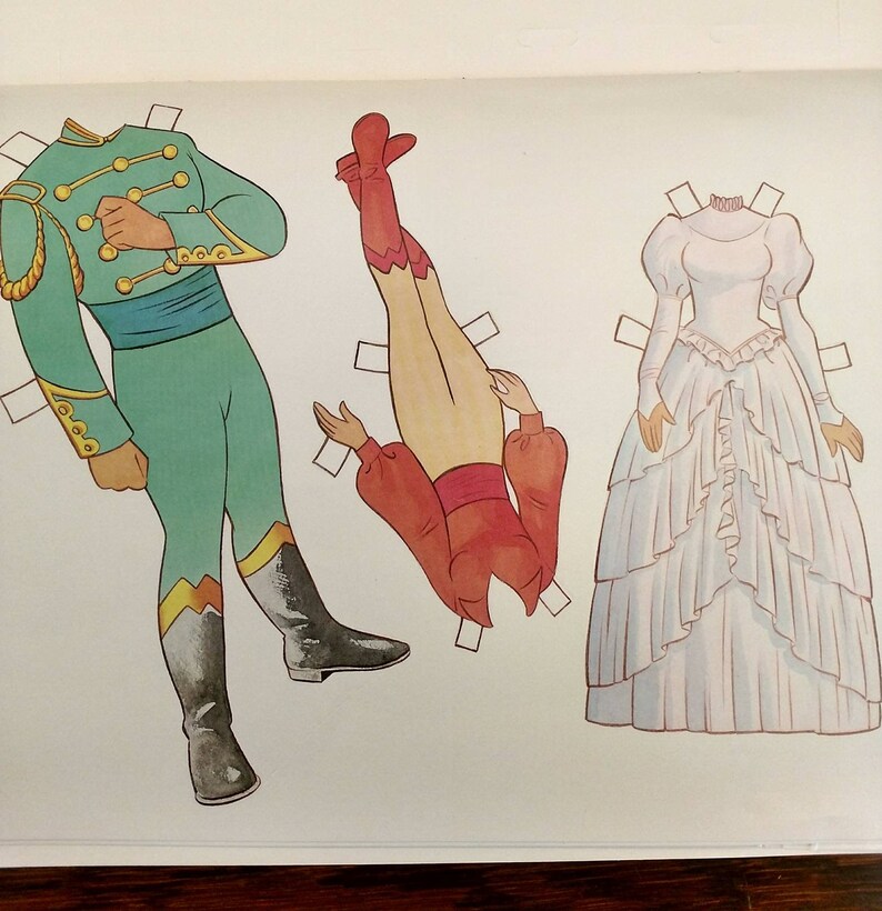 May include: A paper doll set featuring three figures. The first figure is a man wearing a green uniform with gold trim. The second figure is a woman wearing a red and white dress. The third figure is a woman wearing a white dress with a ruffled skirt.