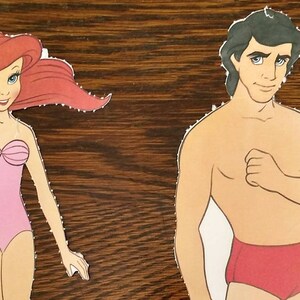 May include: Paper doll cutouts of Ariel and Prince Eric from the Disney film The Little Mermaid. Ariel is wearing a pink swimsuit and has long red hair. Prince Eric is wearing red swimming trunks and has short black hair.
