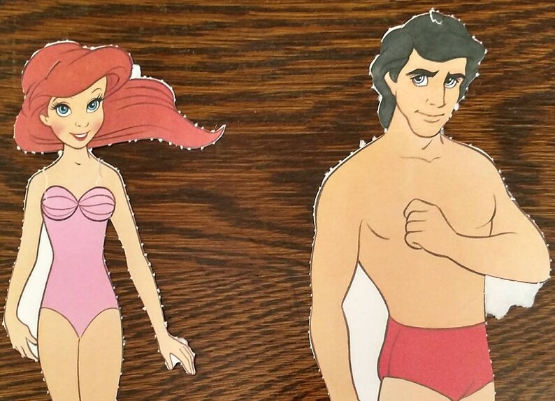 May include: Paper doll cutouts of Ariel and Prince Eric from the Disney film The Little Mermaid. Ariel is wearing a pink swimsuit and has long red hair. Prince Eric is wearing red swimming trunks and has short black hair.
