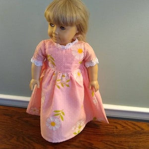 May include: A doll wearing a pink dress with white polka dots and daisy floral print. The dress has short sleeves with white lace trim and a matching white lace collar. The doll has blonde hair and is standing on a wooden surface.