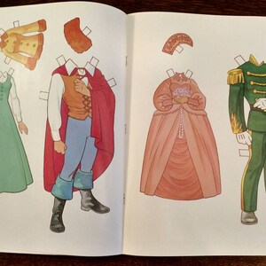 May include: A paper doll set featuring a princess, a prince, and a soldier. The princess wears a pink gown with a white lace trim and a crown. The prince wears a red cape, a blue shirt, and brown trousers. The soldier wears a green uniform with gold trim and a white hat.