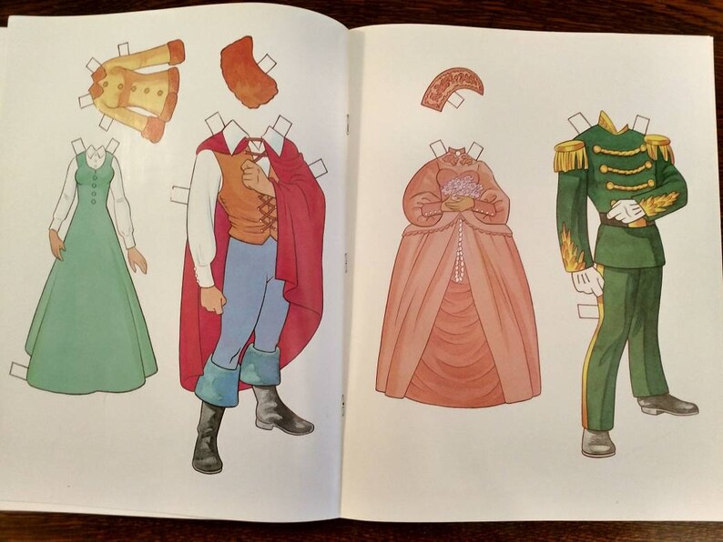 May include: A paper doll set featuring a princess, a prince, and a soldier. The princess wears a pink gown with a white lace trim and a crown. The prince wears a red cape, a blue shirt, and brown trousers. The soldier wears a green uniform with gold trim and a white hat.