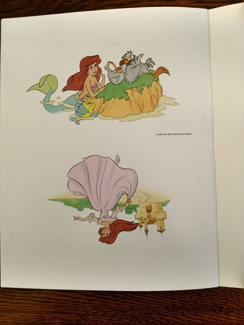 May include: A page from a children's book featuring a mermaid with red hair and a tail, a bird, and a fish. The bottom half of the page shows a woman with red hair wearing a pink dress, upside down, with a castle in the background. The text "&copy;1989 The Walt Disney Company" is at the bottom of the page.