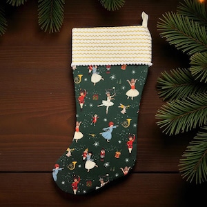 Puede incluir: Un calcetín navideño con un fondo verde oscuro y un estampado de bailarines, músicos y regalos. La parte superior es blanca con líneas onduladas doradas y un ribete blanco. Una presilla blanca está unida en la parte superior.