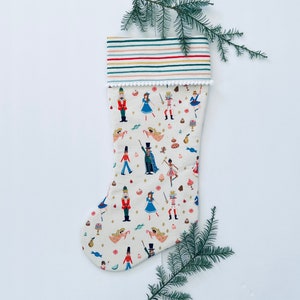 May include: A white Christmas stocking with a Nutcracker theme. The stocking is decorated with a colorful pattern of Nutcracker characters, candy canes, and other holiday elements. The stocking has a white cuff with a scalloped edge.