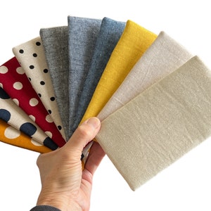 Puede incluir: Un abanico de muestras de tela rectangulares en varios colores y patrones, incluyendo amarillo mostaza con puntos blancos, crema con puntos azul marino, rojo con puntos blancos, crema con puntos negros, gris sólido, azul grisáceo, amarillo mostaza y beige sólido.