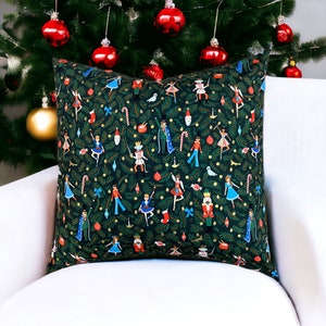 May include: A decorative square pillow with a dark green background featuring a repeating pattern of Christmas-themed illustrations. The pattern includes nutcrackers, dancers, candy canes, and ornaments. The pillow is placed on a white surface.