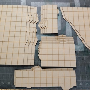 May include: Various laser-cut wooden terrain pieces for tabletop gaming. The pieces are light brown with a grid pattern, and some have irregular edges, suggesting a ruined or broken structure. The pieces are arranged on a dark gray cutting mat.