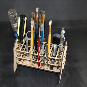 May include: A wooden stand with multiple slots for holding various tools, including paintbrushes, screwdrivers, and a small airbrush. The stand is made of light brown wood and has a rectangular shape.