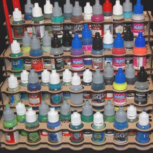May include: A wooden paint bottle organizer filled with various colors of paint bottles. The bottles have different colored caps and labels. The organizer is made of layered wood and has multiple tiers for storage.
