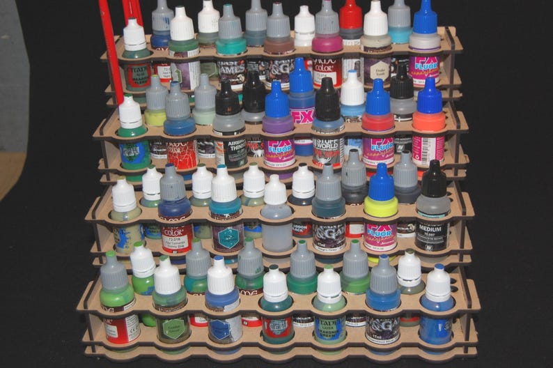 May include: A wooden paint bottle organizer filled with various colors of paint bottles. The bottles have different colored caps and labels. The organizer is made of layered wood and has multiple tiers for storage.