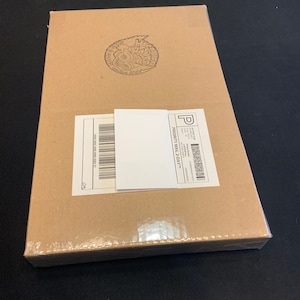 May include: A rectangular cardboard box sealed in clear plastic wrap. A white shipping label and a smaller white label are affixed to the box. A circular stamp is visible on the top of the box.