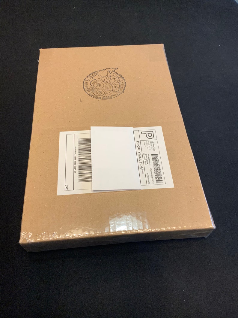 May include: A rectangular cardboard box sealed in clear plastic wrap. A white shipping label and a smaller white label are affixed to the box. A circular stamp is visible on the top of the box.