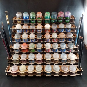 May include: A wooden paint rack filled with various colors of paint pots. The rack is designed to hold multiple pots, with slots for each. Paintbrushes are placed on the sides. The paint colors include shades of blue, green, brown, and purple.