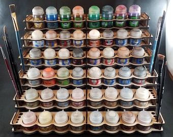 May include: A wooden paint rack filled with various colors of paint pots. The rack is designed to hold multiple pots, with slots for each. Paintbrushes are placed on the sides. The paint colors include shades of blue, green, brown, and purple.