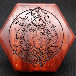 May include: A wooden hexagonal box with a circular design featuring a detailed engraving of a hooded figure with an eyepatch. The figure has a serious expression and is wearing a cloak with intricate details.