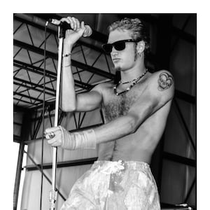 May include: Black and white image of a person on stage holding a microphone. The person is wearing sunglasses, a necklace, and shorts. A tattoo of a skull is visible on the person's shoulder. The person is holding a microphone stand with a wrapped wrist.