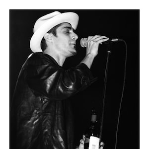 May include: A man in a white fedora and a black leather jacket sings into a microphone while holding a bottle of wine.