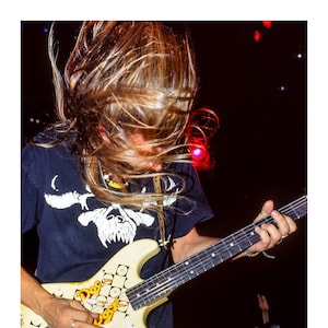 May include: A musician with long blonde hair plays a white electric guitar with black and yellow graphics. The guitar has the words "Dog Rock" written on it.