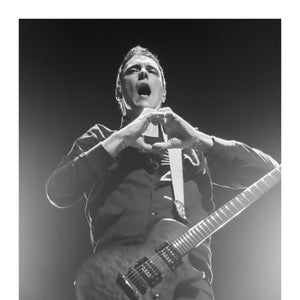 May include: A black and white photo of a man singing into a microphone while holding a black electric guitar. He is making a heart shape with his hands.