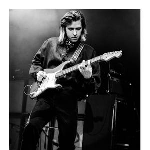 May include: Black and white image of a person playing an electric guitar on stage. The guitar is a classic design with a white pickguard and a light-colored body. The person is wearing a dark shirt and pants, with a patterned scarf around the neck.