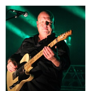 May include: A man playing a light brown electric guitar with a black pickguard. He is wearing a black shirt and is standing on a stage with green lights.