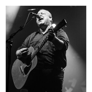 May include: A man in a black shirt sings into a microphone while playing an acoustic guitar. He is on stage with a spotlight shining on him.