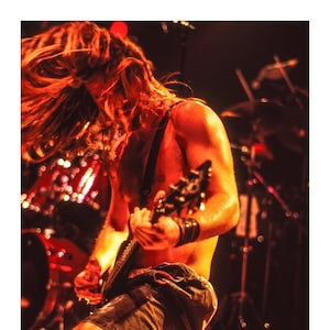 May include: A man with long, flowing red hair plays an electric guitar on stage. He is wearing a black tank top and camouflage shorts. The stage is lit with warm, orange light.
