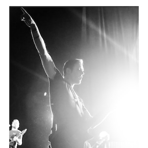 May include: A musician performs on stage, holding a guitar and singing into a microphone. The stage is lit with bright spotlights, creating a dramatic effect.