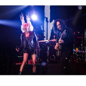 May include: A woman with blonde hair wearing a black leather jacket and fishnet stockings sings into a microphone while a man with long dark hair plays a guitar on stage.
