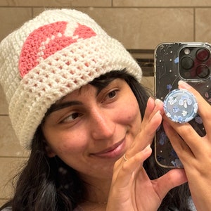 May include: A white crocheted beanie with a pink crocheted flower design. The beanie is worn by a person holding a phone case with a floral design.