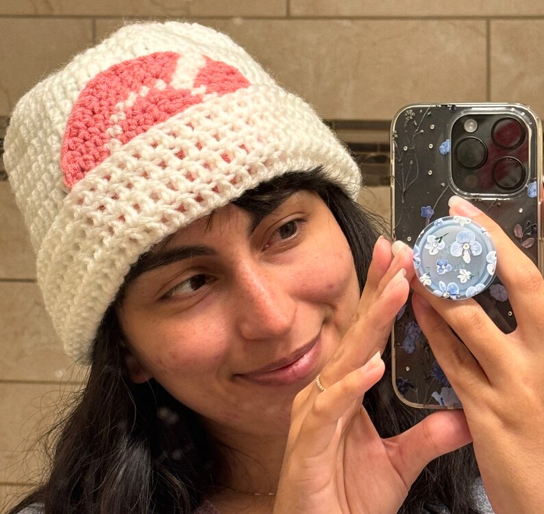 May include: A white crocheted beanie with a pink crocheted flower design. The beanie is worn by a person holding a phone case with a floral design.