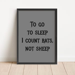 Può includere: Stampa incorniciata con il testo "To go to sleep I count bats, not sheep" in carattere gotico nero su sfondo grigio. La cornice è nera e la stampa è esposta contro un muro bianco.