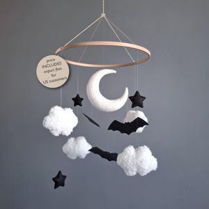 May include: A whimsical baby mobile featuring a wooden ring with hanging felt shapes. The mobile includes a white crescent moon, black stars, a black bat, and fluffy white clouds. The mobile is suspended by a thin string against a gray background.