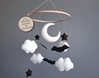 May include: A whimsical baby mobile featuring a wooden ring with hanging felt shapes. The mobile includes a white crescent moon, black stars, a black bat, and fluffy white clouds. The mobile is suspended by a thin string against a gray background.