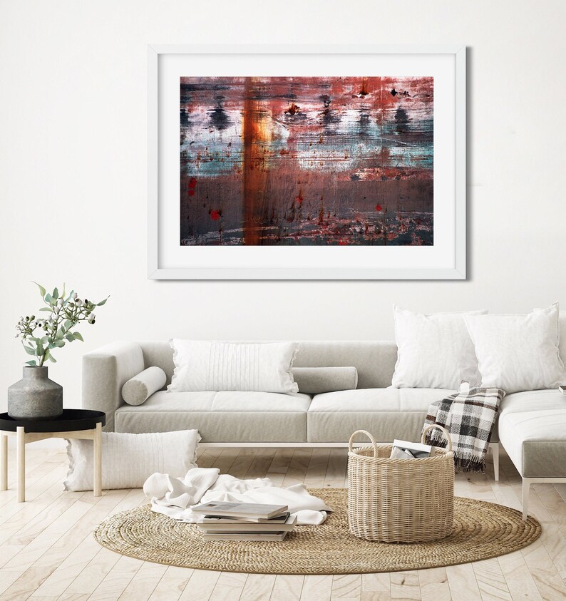 May include: A framed abstract painting with a white frame. The painting features a variety of colors, including red, orange, blue, and green. The painting is hung on a white wall above a white couch with pillows and a throw blanket.