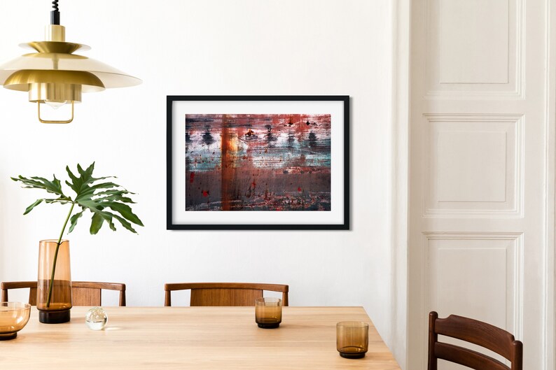 May include: Abstract painting in a black frame with red, orange, blue, and white colors. The painting is hanging on a white wall above a wooden table.