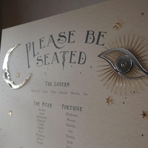 May include: A beige seating chart sign with the text "PLEASE BE SEATED" in dark gray lettering. It features a silver crescent moon on the left, a silver eye icon with rays on the right, and small gold star accents. Names are listed under headings.