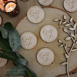 May include: Wooden save the date magnets featuring the text "Save the Date Scott & Jo 22.09.2018" in a cursive script. The magnets are circular and have a natural wood finish.