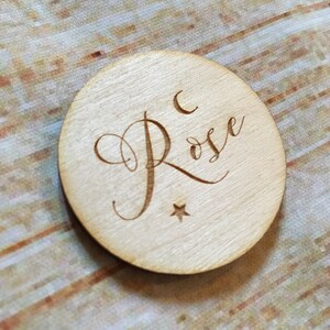 May include: A round wooden disc with the name "Rose" engraved in cursive script. A crescent moon and a star are also engraved on the disc.