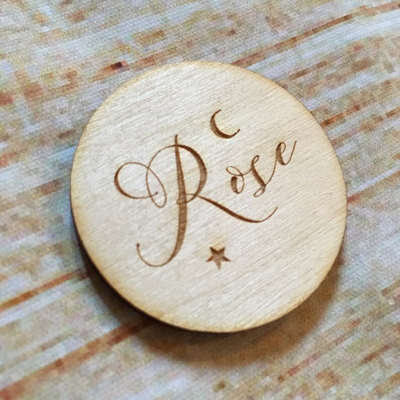 May include: A round wooden disc with the name "Rose" engraved in cursive script. A crescent moon and a star are also engraved on the disc.