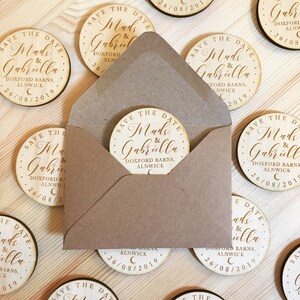 May include: A set of round wooden save the date cards with the text "Save the Date" and the names "Mads & Gabriella" in cursive script. The cards also include the date "26/08/2019" and the location "Doxford Barns, Alnwick".