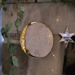 Puede incluir: Una decoración de pared redonda en forma de luna blanca con un borde dorado, un diseño de luna creciente y una pequeña imagen de dos animales en su interior. La luna está colgada en una pared con una cuerda y tiene un espejo en forma de estrella colgado a su lado.