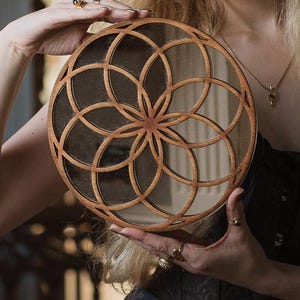 May include: A wooden circular mirror with a geometric design. The mirror is made of wood and has a dark brown finish. The design is a flower-like pattern with a circle in the center.