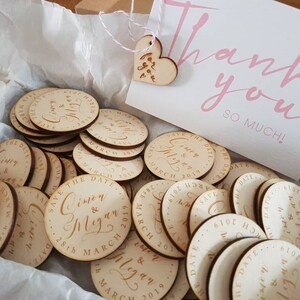 May include: A box filled with round wooden save the date magnets featuring the names Simon & Megan and the date 28th March 2019. The box also includes a small wooden heart with the text "Thank you so much!" written on it.