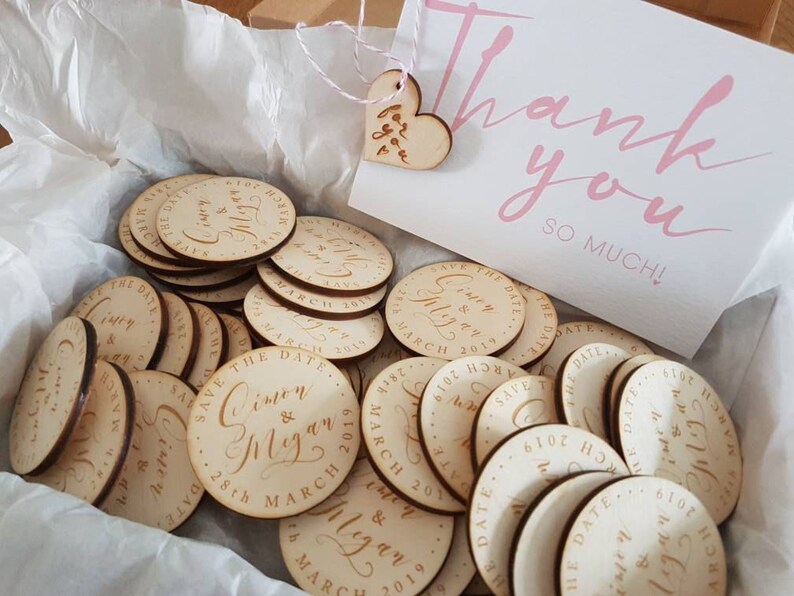 May include: A box filled with round wooden save the date magnets featuring the names Simon & Megan and the date 28th March 2019. The box also includes a small wooden heart with the text "Thank you so much!" written on it.