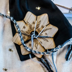 May include: A wooden star-shaped craft with a sun and star design, with a braided cord and various strands of yarn in shades of white, grey, and black. A black bag and gold star charms are also present.