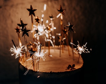 Op de afbeelding: Een witte taart met gouden stervormige kaarsen en brandende vuurwerk op de bovenkant. De taart staat op een donkere achtergrond.