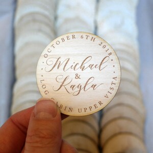May include: Round wooden wedding favour with the text "October 6th 2018...Log Cabin Upper Vista...Michael & Kayla" engraved in brown.