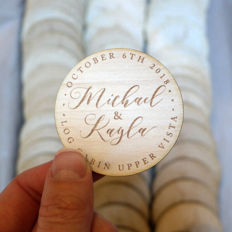 May include: Round wooden wedding favour with the text "October 6th 2018...Log Cabin Upper Vista...Michael & Kayla" engraved in brown.
